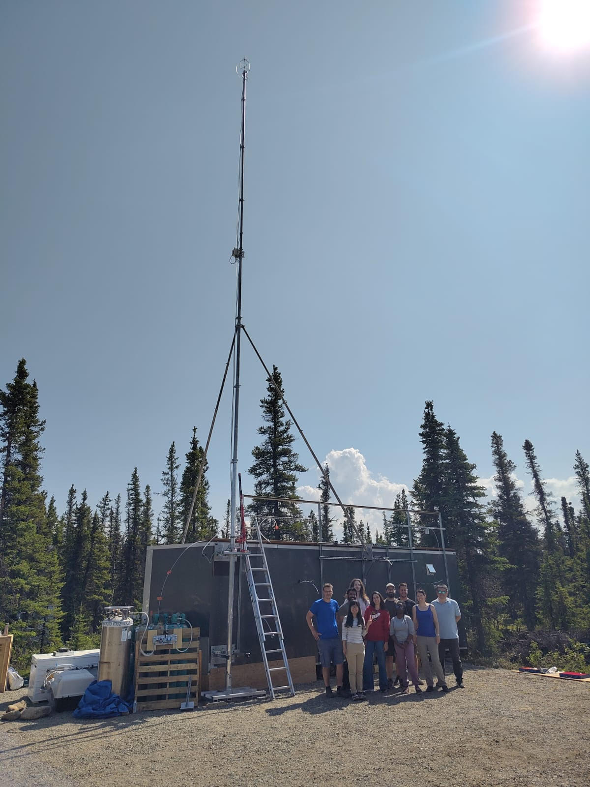 Group poses for a photo infront of their air monitoring equipment in a flat gravel area with pine trees behind them on a cloudless summer day