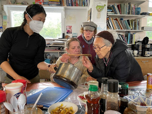 Four women gather around a silver pot. One woman offers a wooden spoon to another for testing. 