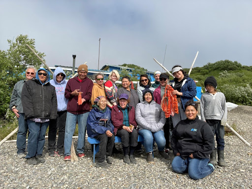 A group of people pose on a gravel beach. Some are holding strips of salmon filets. 