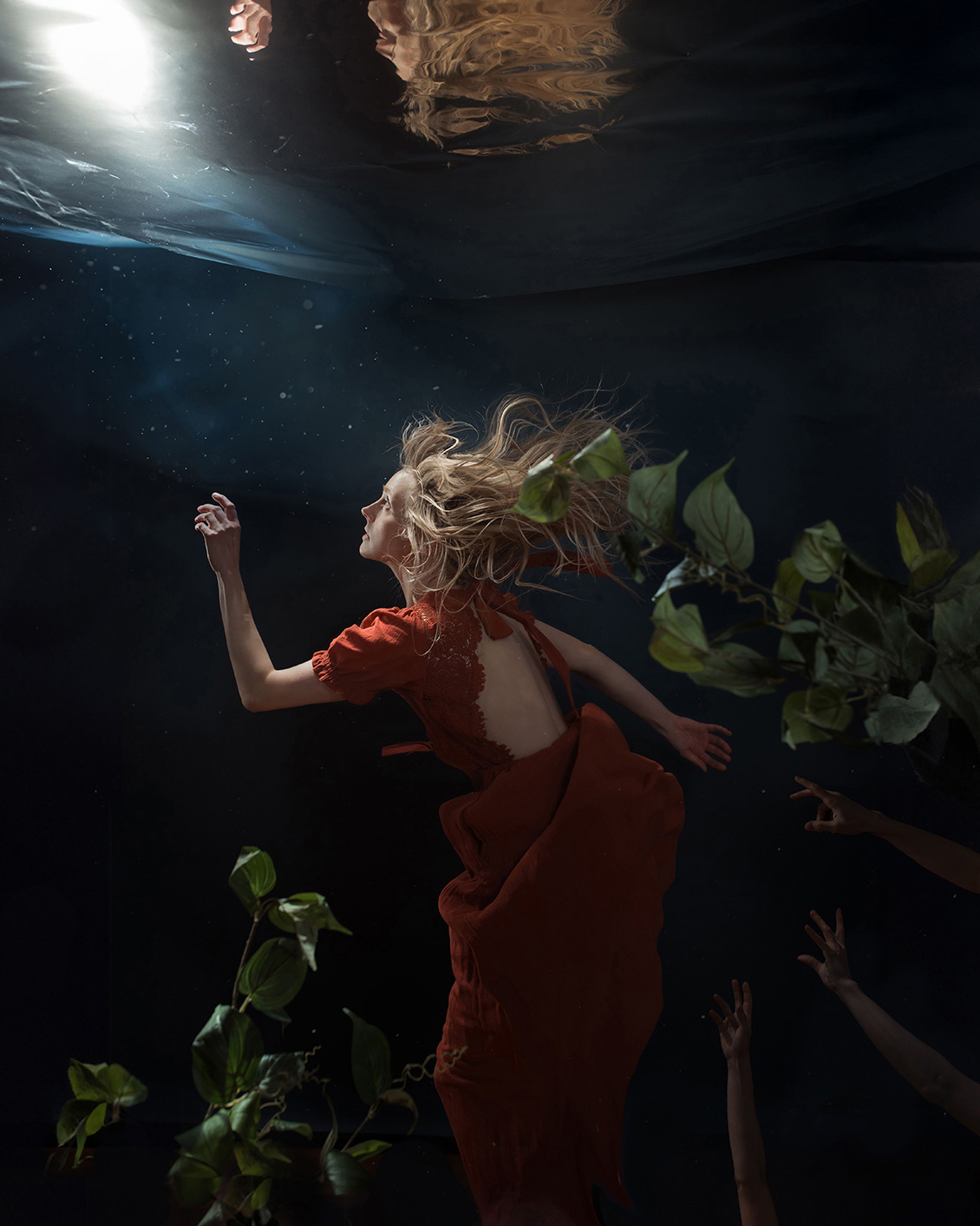Woman in a red dress underwater with a hand extended upwards towards light. Arms reach towards her from the bottom right