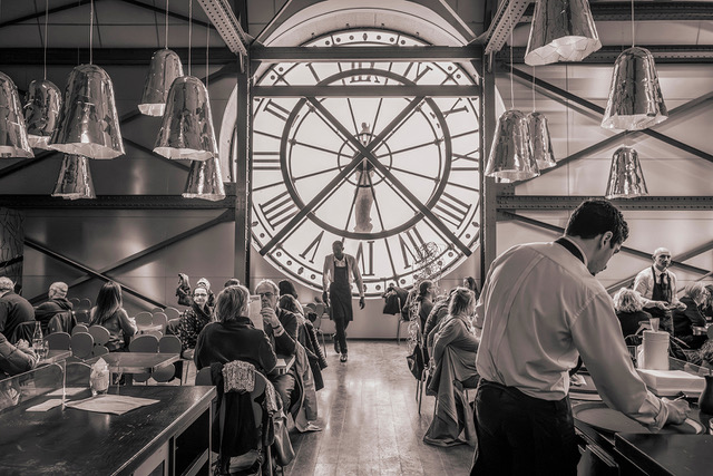 Black and white photo of a cafe in Paris. A large clock face is in the background. Photo by Charles Mason
