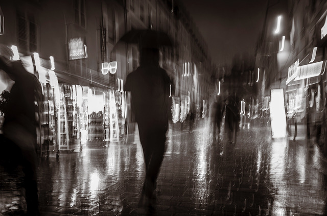Black and white photo of people walking through the streets of Paris with umbrellas at night. Photo by Charles Mason