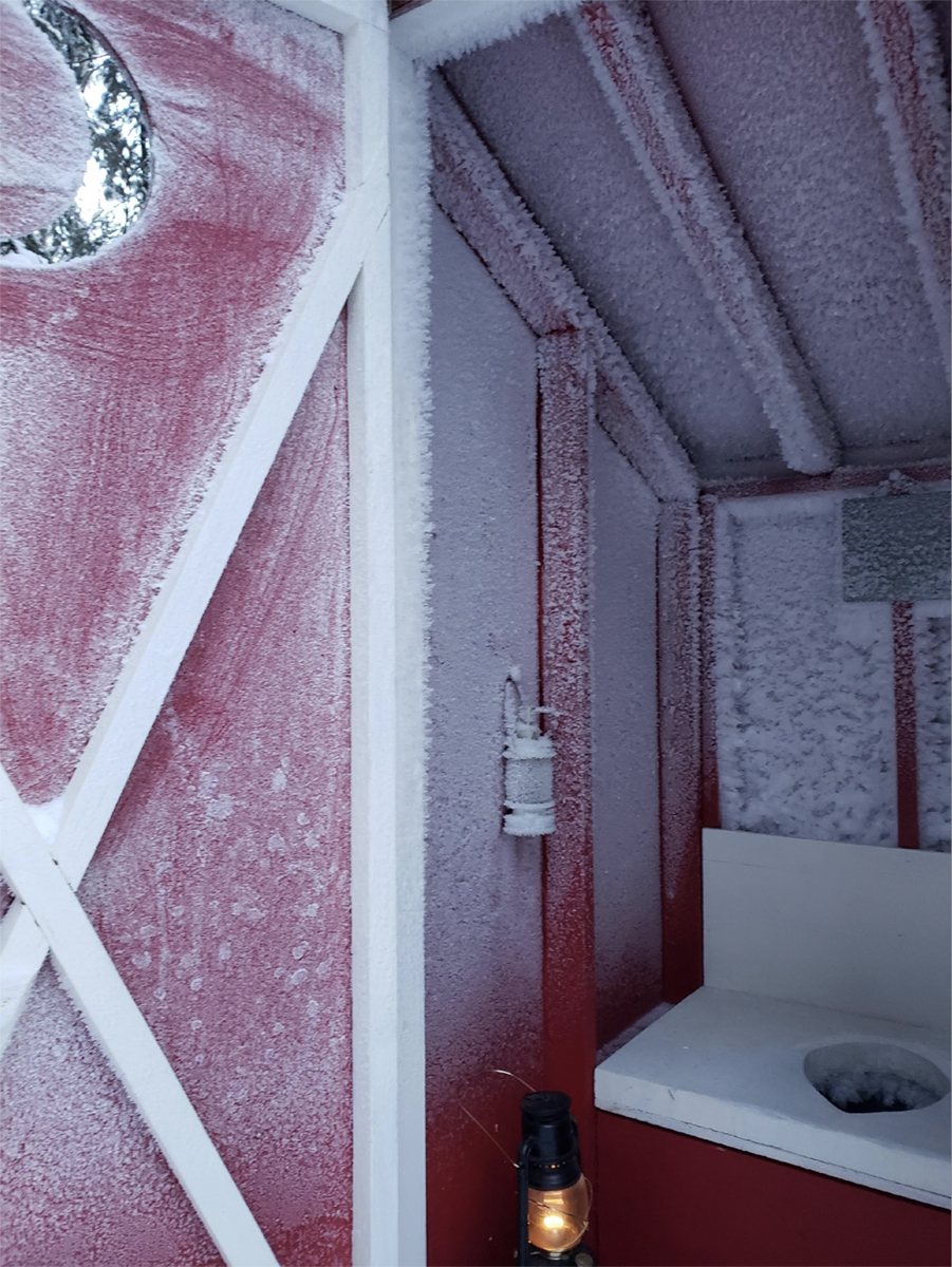 A frozen outhouse with the door ajar. The walls are red and covered in ice. Photo courtesy of Arciniega