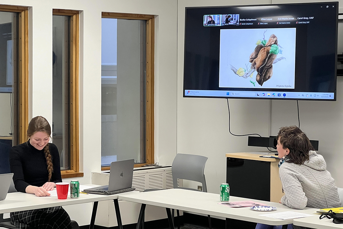 Samantha Botz (left) smiles while looking at her paper. The TV screen shows an illustration of a mushroom and a student looks on. UAF Photo by Carol Gray.