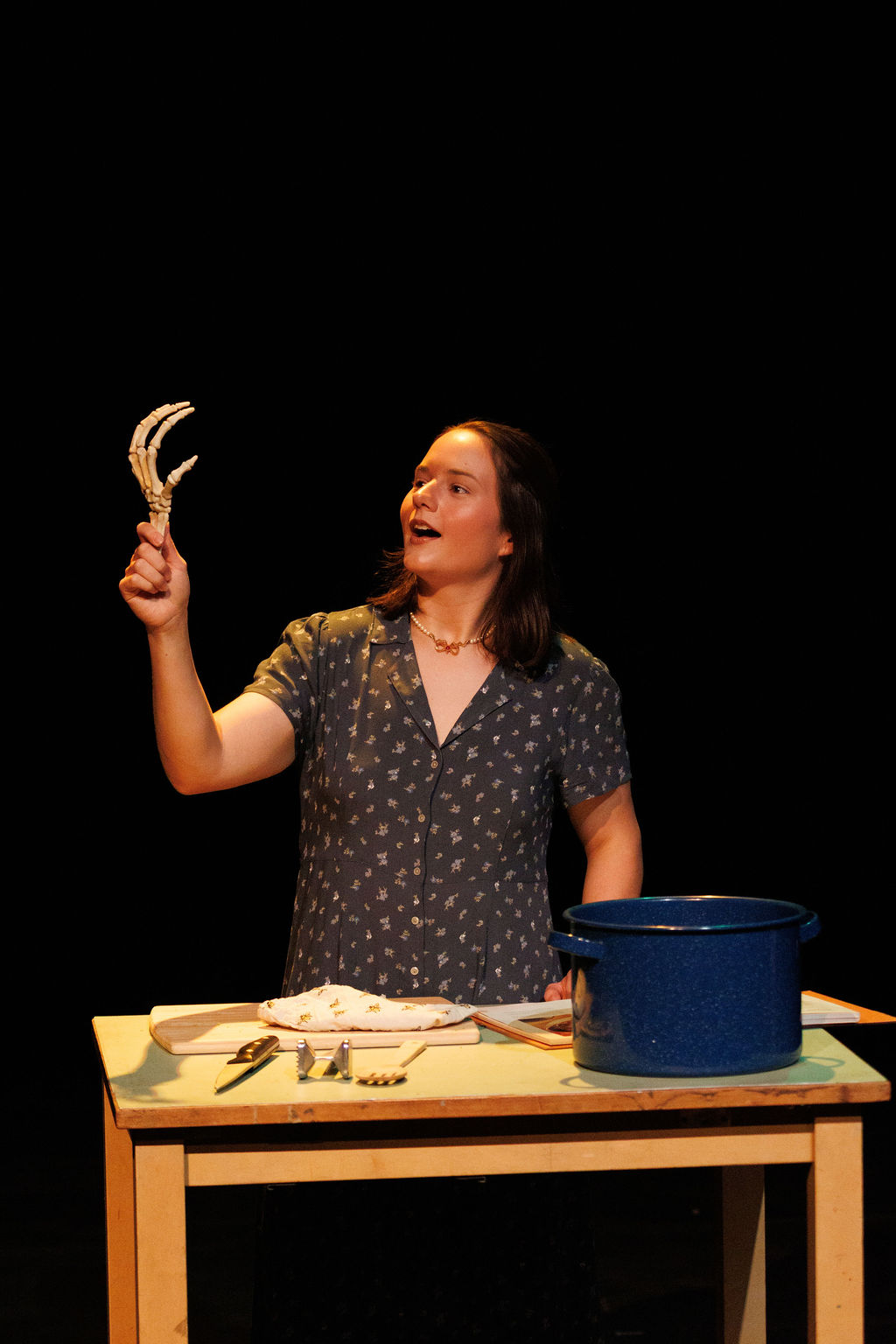 Deyanira Van Acker, as Mary, holds up a chicken foot over a table and soup pot.