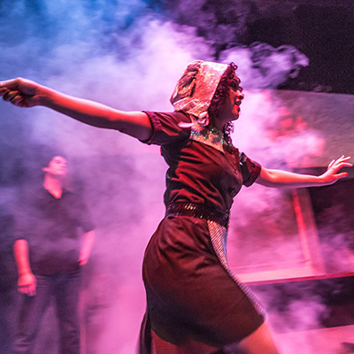A theatre student in costume gestures while moving across a foggy neon-lit stage