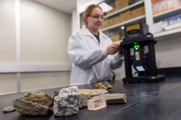 A table with rock samples and a woman in a white coat adjusting a microscope