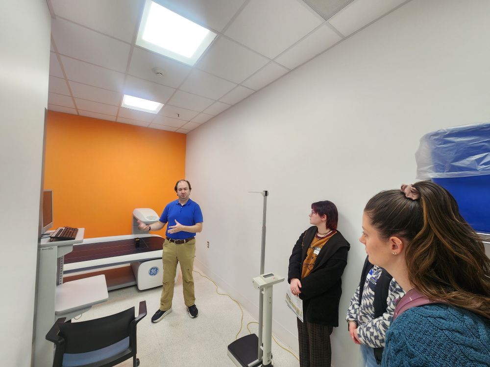 Man in blue polo shirt explains the functions of a table top x-ray machine to three people in a medical looking office space with an orange accent wall. 