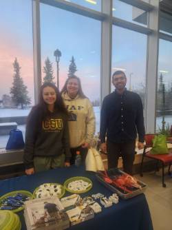 Three people stand behind a table full of veterinary medicine program materials and swag in a room with floor to ceiling windows. A view of a cold winter sunrise and pine trees outside.