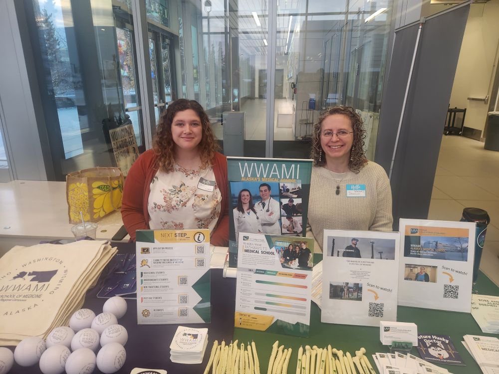 Two women with curly brown hair sit smiling behind a table full of WWAMI program promotional items in a brightly lit room with a glass entryway behind them. 