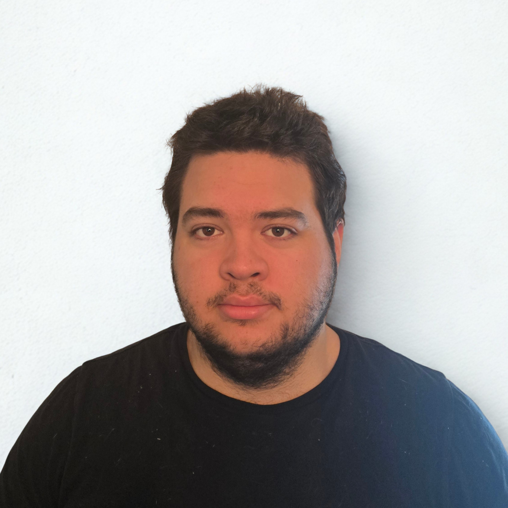 A head-and-shoulders portrait of a young man with short, dark, textured hair and a light beard. He is wearing a plain black t-shirt and looking directly at the camera with a neutral expression. He is positioned against a clean, solid white background.