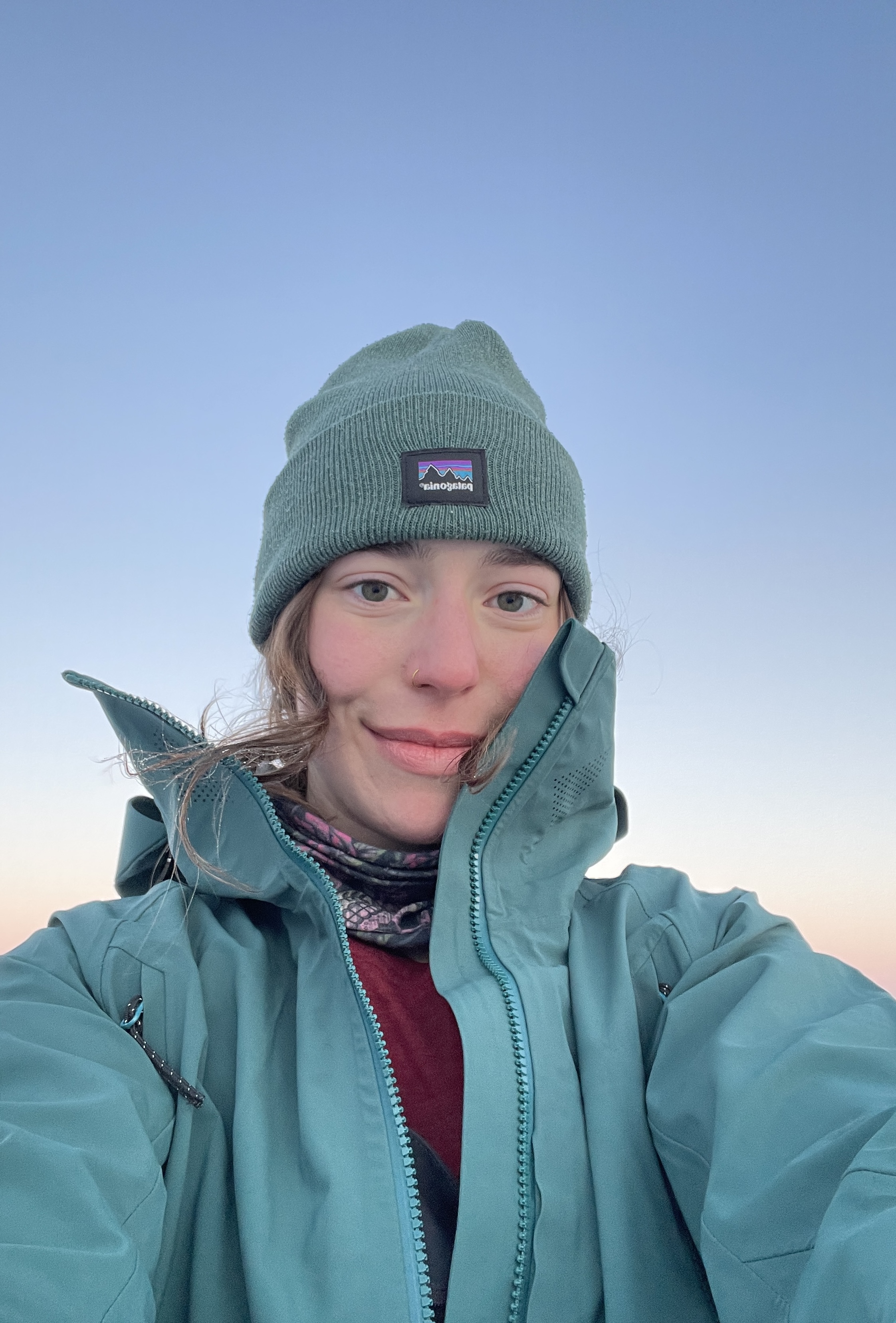 A selfie of a smiling person outdoors, dressed in winter gear. They are wearing a sage green Patagonia beanie and a matching teal technical jacket zipped up to their chin. A few strands of brown hair are windswept across their face against a soft, gradient sky that transitions from pale blue to a warm sunset pink.