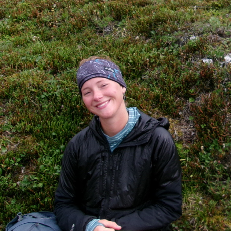 A person sits on a grassy, wildflower-covered hillside, smiling warmly. They are wearing a black hooded jacket over a striped teal shirt and a patterned purple and blue headband. A grey hiking backpack is visible at their side.