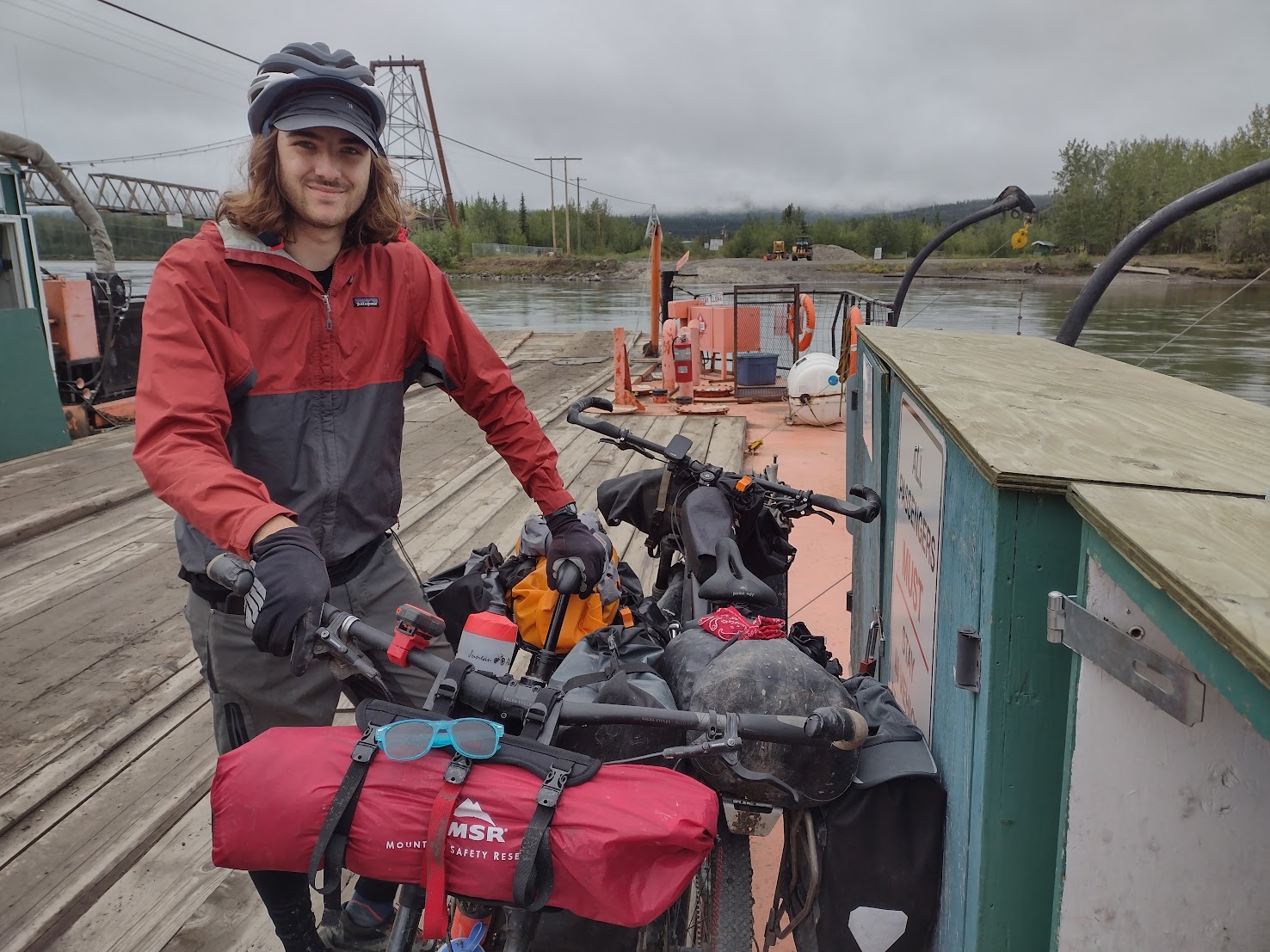 A person with long brown hair and a bicycle helmet stands on a wooden ferry deck, holding onto their fully loaded touring bike. They are wearing a red and dark grey Patagonia jacket. The bike is equipped with several bags, including a prominent red MSR dry bag on the front handlebar rack. In the background, a wide river flows beneath a grey, overcast sky, with a suspension bridge and forested shoreline visible in the distance.