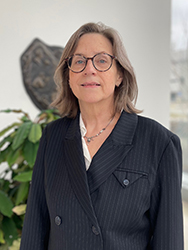 A professional headshot of a woman with shoulder-length light brown hair and dark-rimmed glasses. She is wearing a pinstriped navy blue blazer over a white shirt and a delicate necklace. She is looking directly at the camera with a neutral expression. The background is bright and out of focus, with a green leafy plant visible on the left.