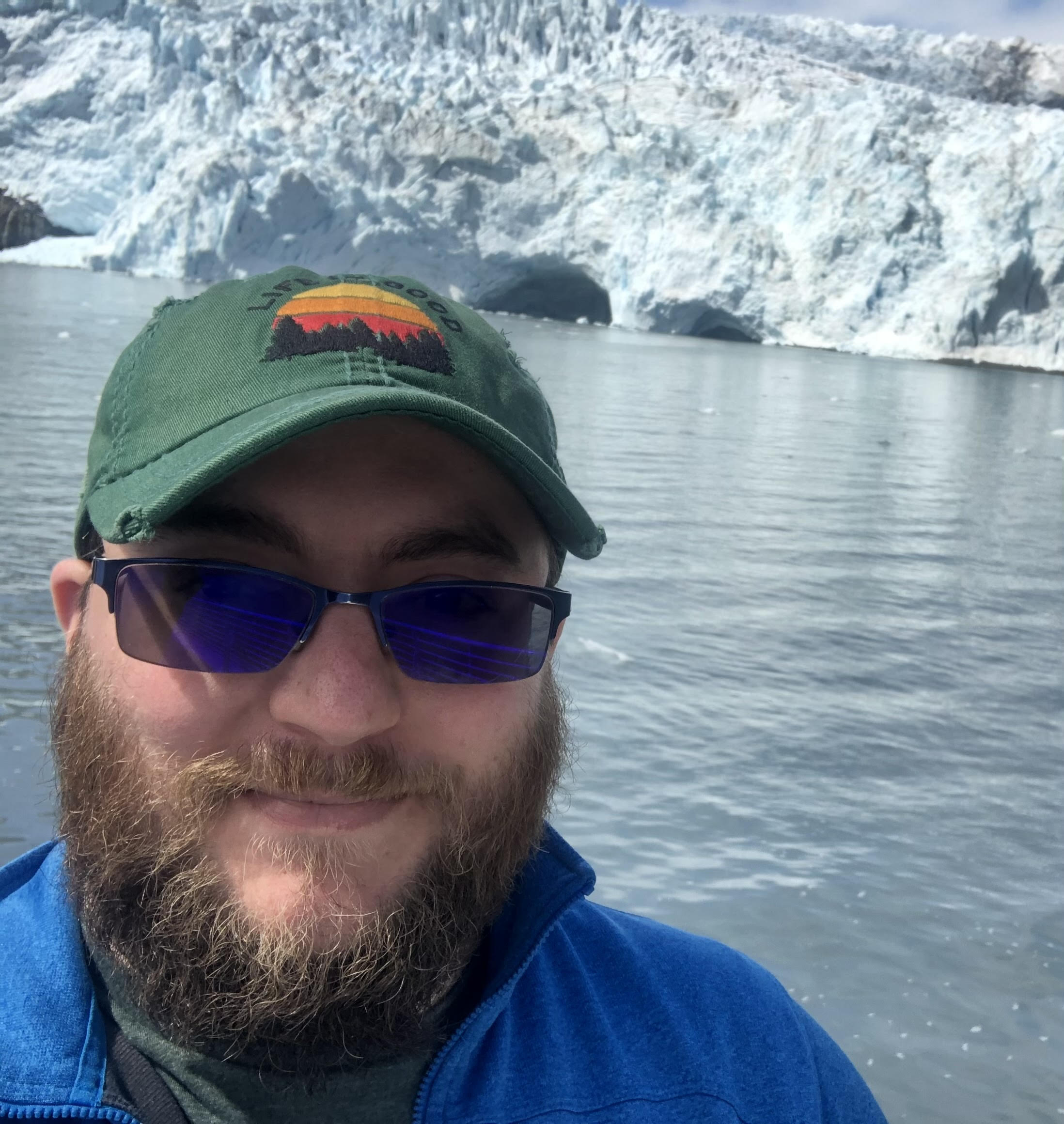 A selfie of a smiling man with a brown beard and blue-tinted sunglasses. He is wearing a bright blue fleece and a green baseball cap featuring a "Life is Good" sunset mountain logo. In the background, a massive, jagged blue glacier meets the water's edge under a clear sky.