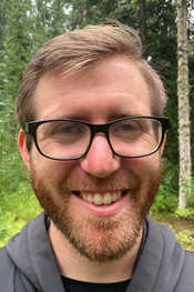 A close-up portrait of a smiling man with short brown hair, a reddish-brown beard, and black-rimmed glasses. He is wearing a grey zip-up hoodie over a black shirt. The background is a soft-focus lush green forest.