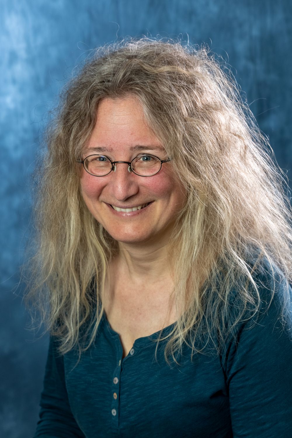 A head-and-shoulders portrait of a person with long, voluminous, light-brown frizzy hair and small, round-rimmed glasses. They are smiling warmly at the camera, wearing a dark teal, button-down henley shirt. The background is a textured, mottled blue studio backdrop.