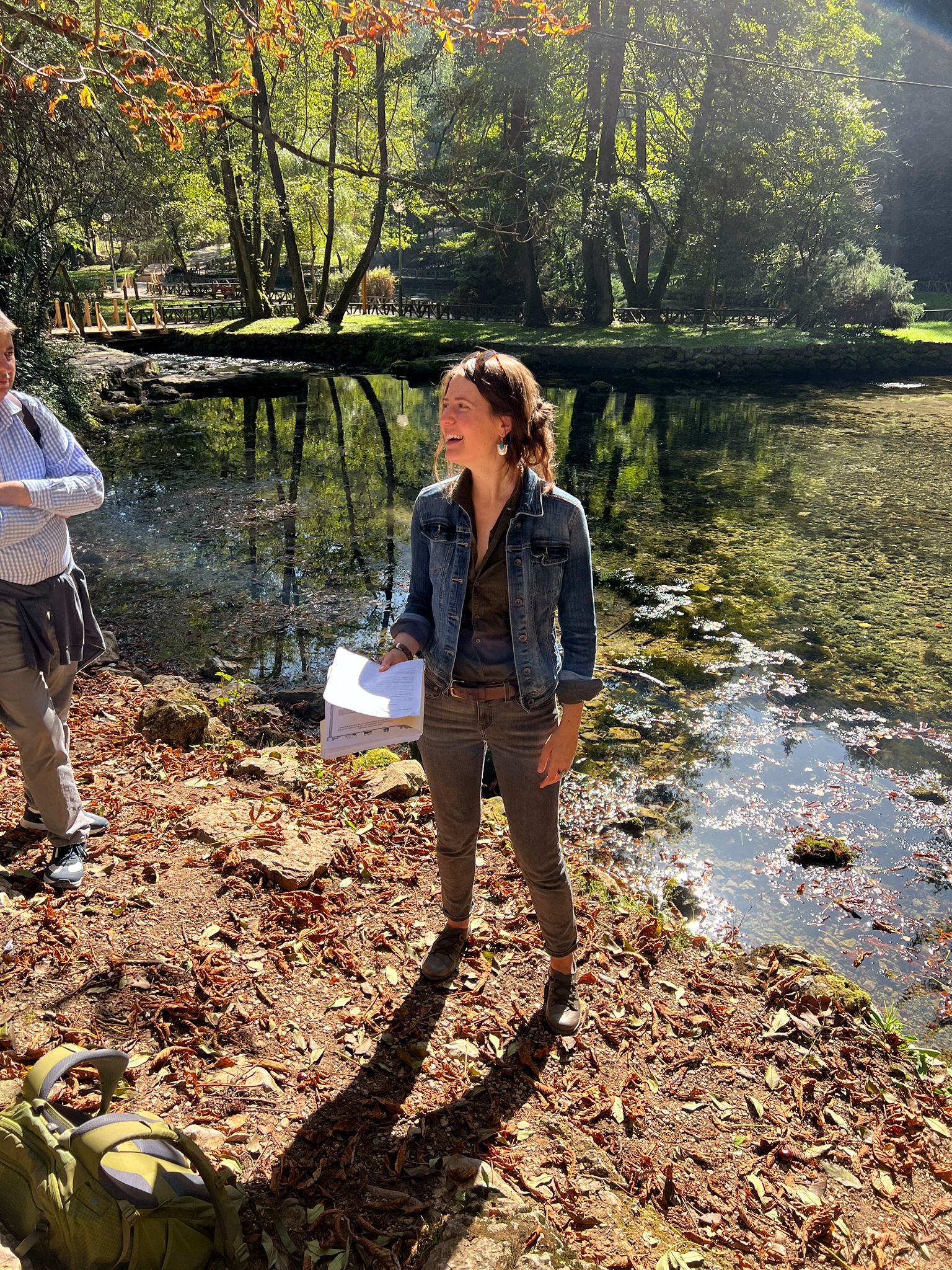 A woman with brown hair tied back stands on a leaf-strewn bank, smiling and looking off-camera. She is wearing a denim jacket over a dark shirt and grey pants, holding a stack of papers. Behind her is a clear, shallow pond reflecting the surrounding lush green trees and bright sunlight. Another person is partially visible on the left, and a green backpack rests on the ground in the foreground.