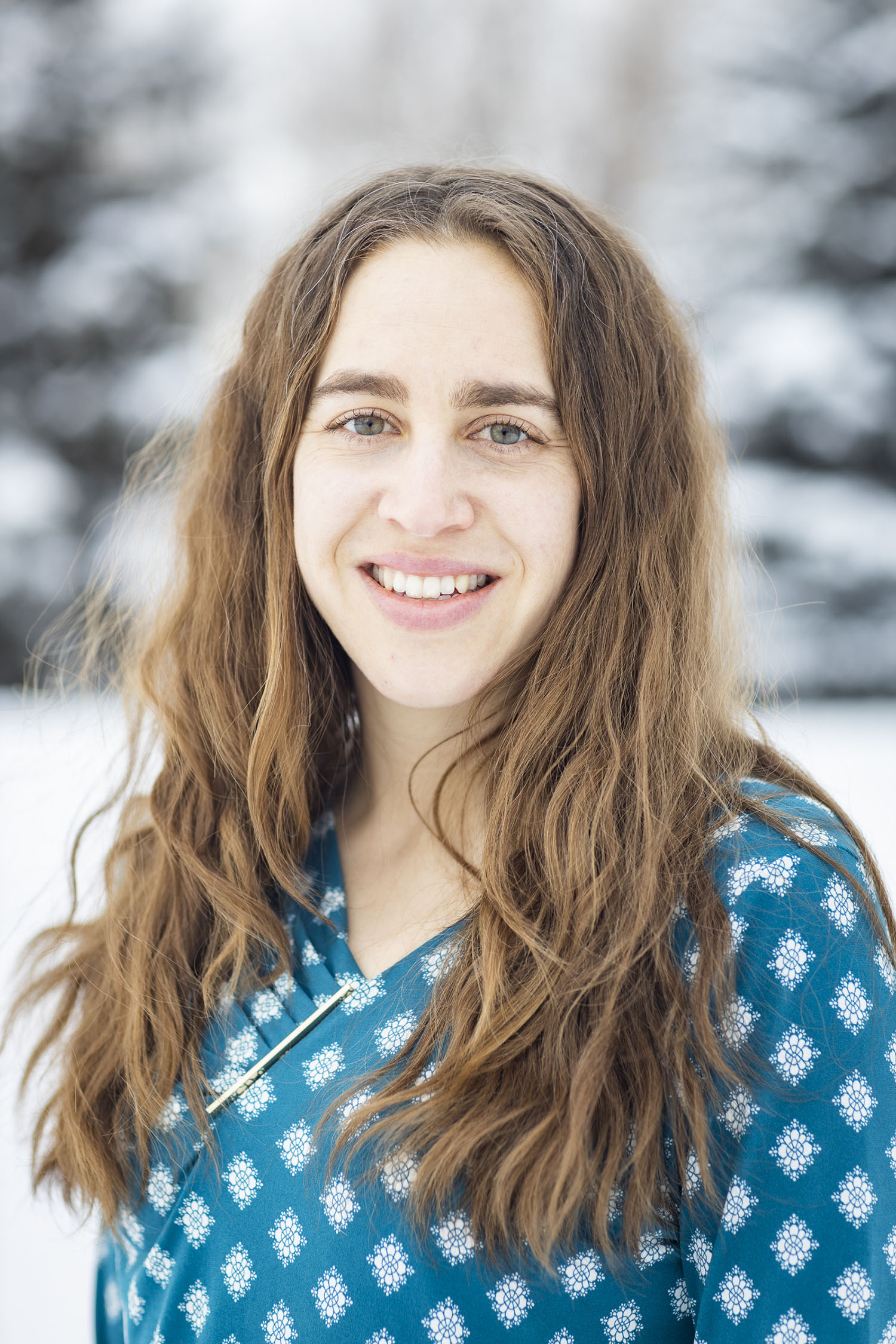 A bright head-and-shoulders portrait of a smiling woman with long, wavy brown hair. She is wearing a teal-colored top with a white geometric pattern and a gold bar pin. The background is a soft-focus winter scene with snow-covered trees.