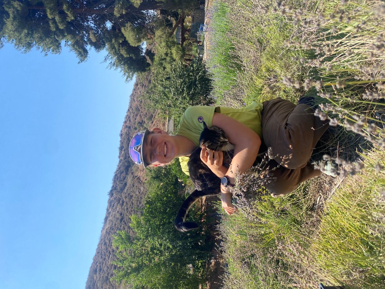 A smiling person crouches in a field of lavender, hugging a dark tabby cat. They are wearing a lime green t-shirt, brown pants, and a grey cap with purple sunglasses perched on top. The background features sun-drenched hills and green trees under a clear blue sky.