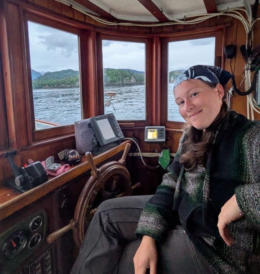 A person with a gentle smile sits in the wood-paneled wheelhouse of a boat. They are wearing a patterned green and black sweater and a dark floral bandana. Beside them is a traditional wooden ship's wheel, various navigation monitors, and controls. Through the large front windows, a calm body of water is visible, surrounded by lush, green, mountainous islands under a cloudy sky.