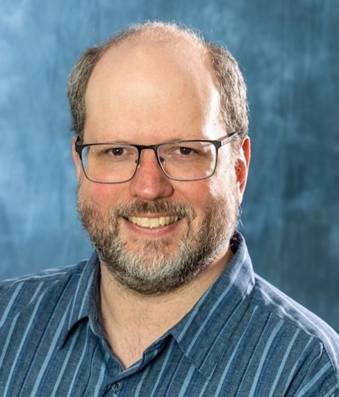 Headshot of a middle-aged man with fair skin, a receding hairline, and a salt-and-pepper beard. He is wearing dark-rimmed rectangular glasses and a blue button-down shirt with thin, vertical white stripes. He is smiling warmly against a textured, mottled blue background.