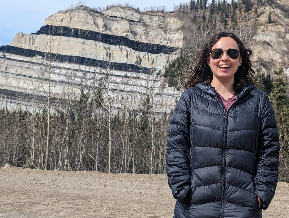 A person with long, dark curly hair and aviator sunglasses smiles brightly while standing in front of a striking geological formation. They are wearing a dark blue puffer jacket over a maroon shirt. In the background, a large, light-colored cliff displays prominent, horizontal black stripes of coal or mineral deposits. A line of bare trees sits at the base of the cliff under a clear sky.