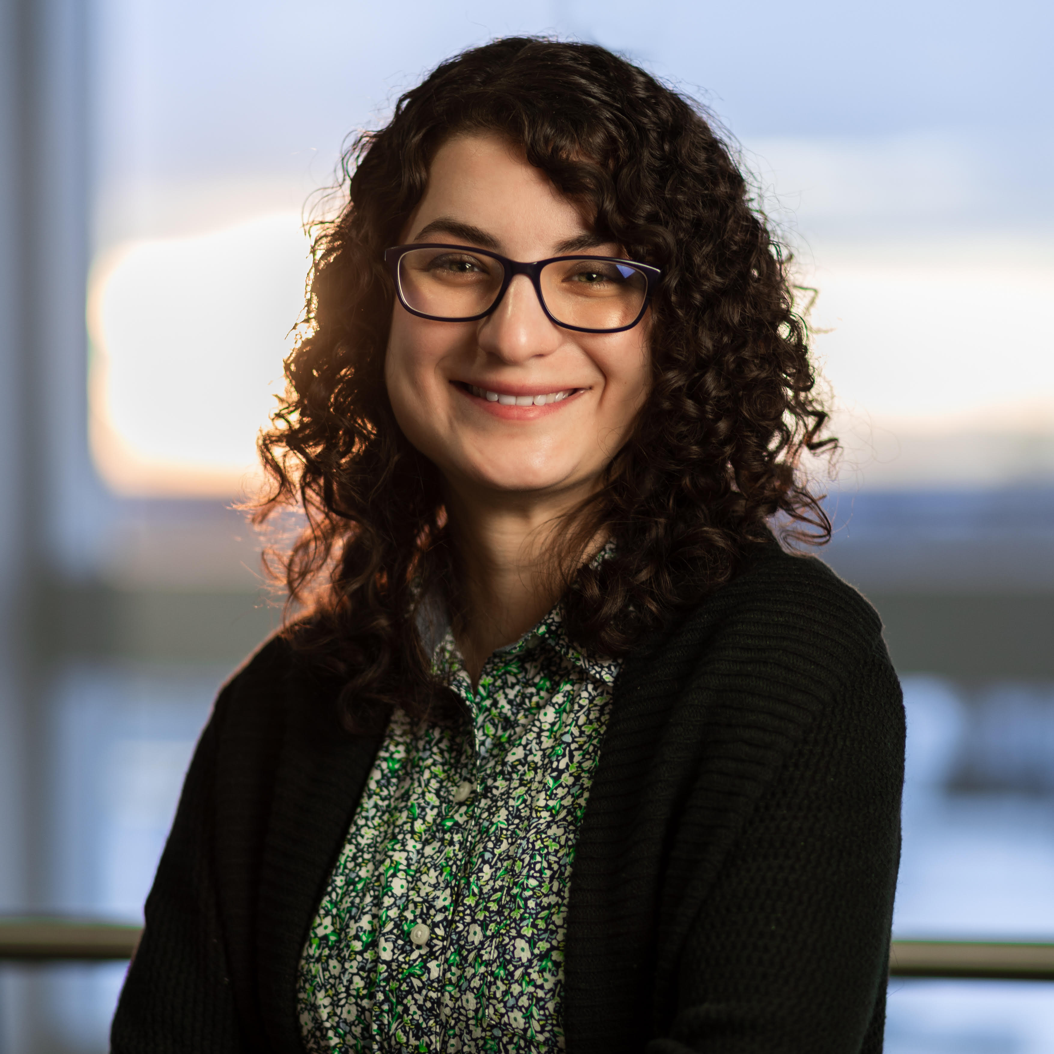 A professional headshot of a smiling woman with dark, curly hair and glasses. She is wearing a green patterned blouse under a black cardigan. The background is a soft-focus indoor setting with a large window showing a view of a city or harbor at dusk.
