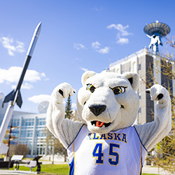 Nook, UAF Mascot, posing in front of West Ridge buildings