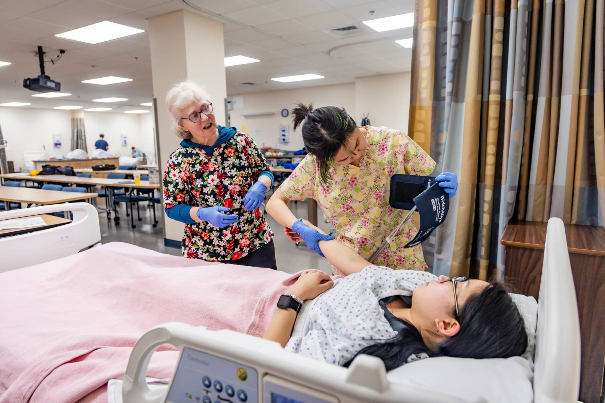 Two nursing students examine a patient lying in a hospital bed.