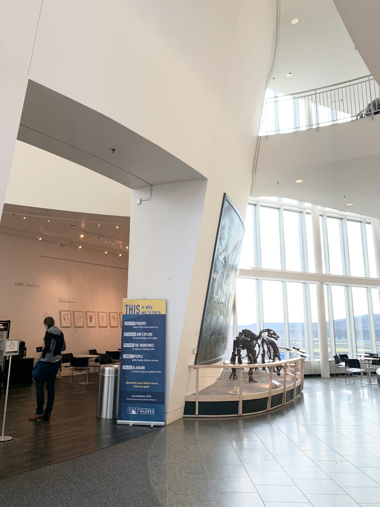 Picture of the cafe space and the lobby overlooking the Tanana Valley and Alaska Range. A woman stands in line at the cafe kiosk. Two dinosaur skeletons are in the lobby in a small display area.