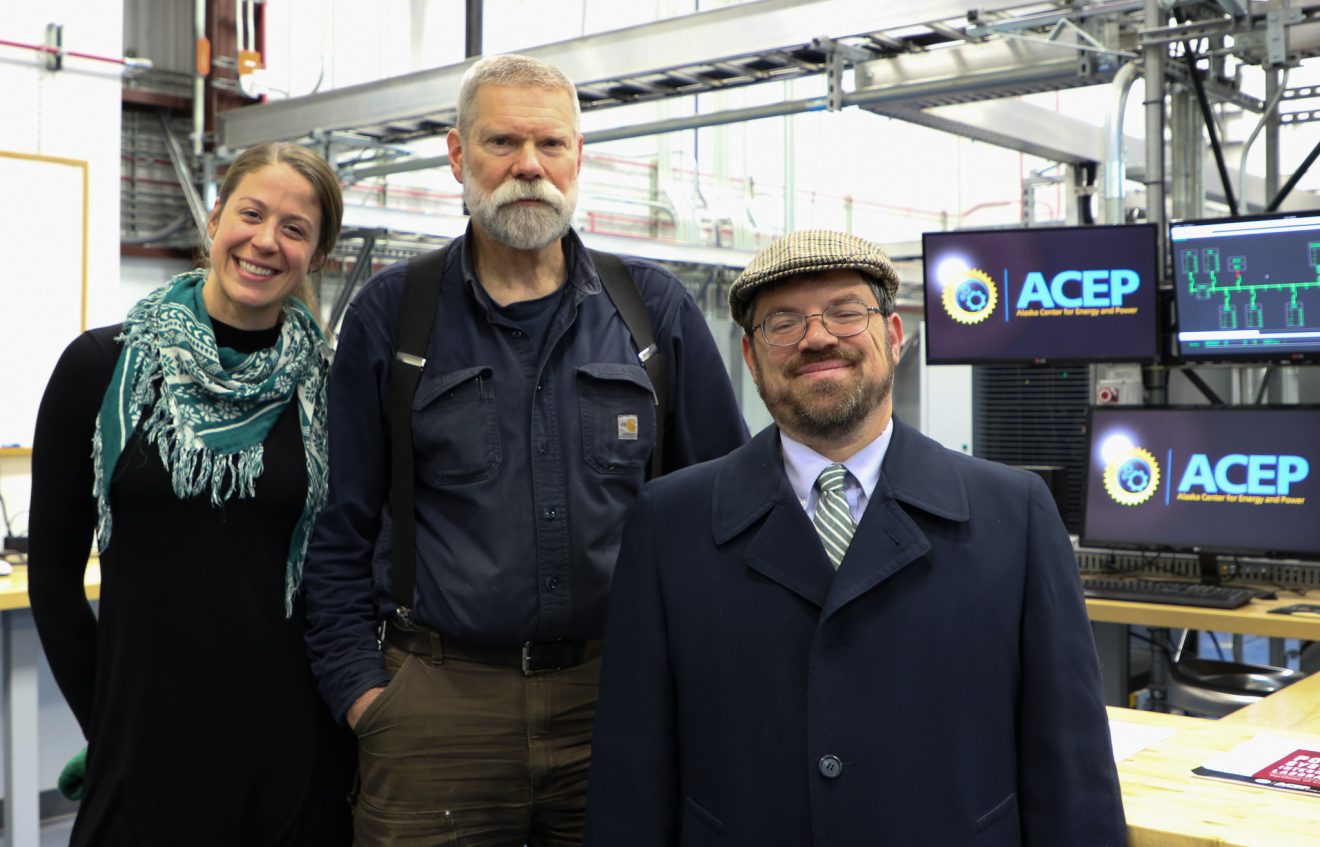 Three people pose in front of three computer screens and some mechanical equipment.