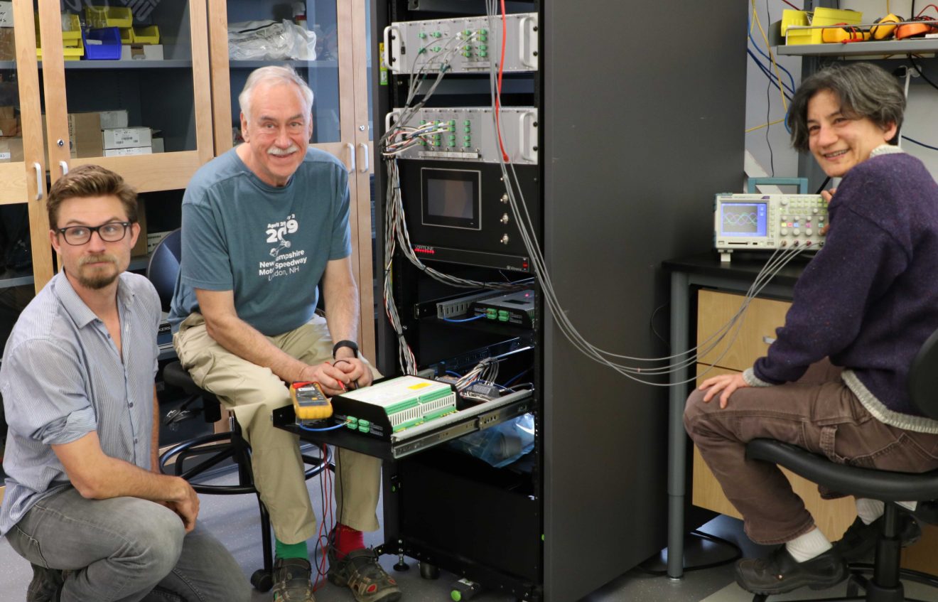 Three people sit in a room filled with computers, cables and wires.