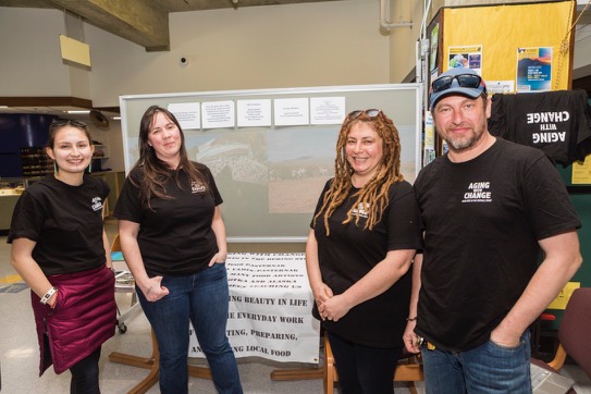 Three women and one man stand in front of a table and large sign as part of a display. It is unclear what the display is about.