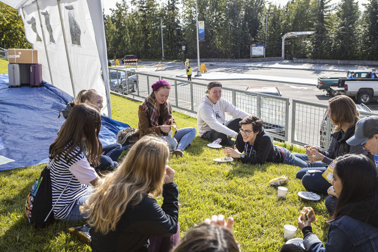 Ten students sit or lie on the grass in a circle outside the MBS complex. Many of them are eating or drinking. Many are smiling or engaging with each other.