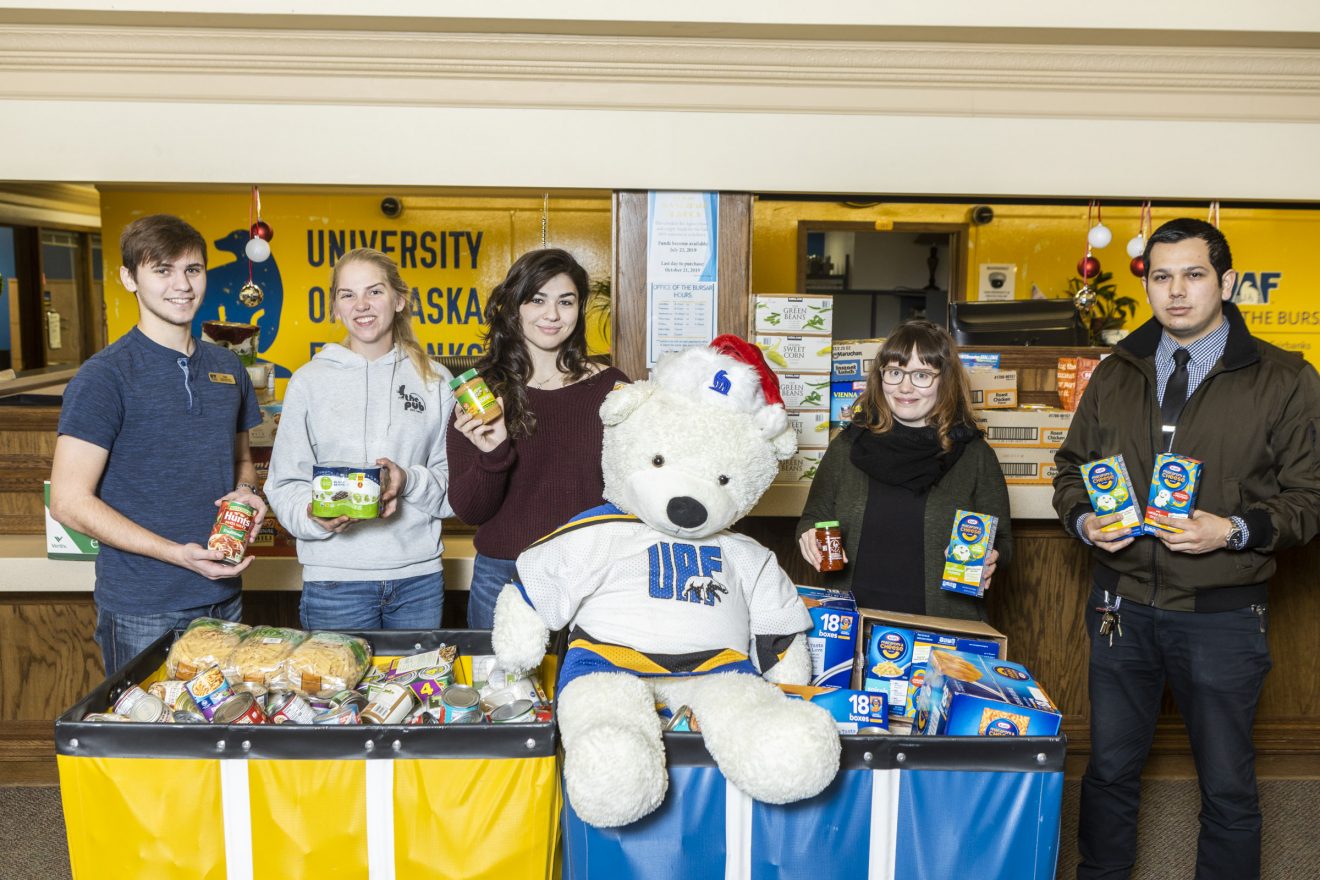 Five people stand behind or beside two large containers filled with food. A stuffed Nanook toy with a Santa hat sits on top of the containers.