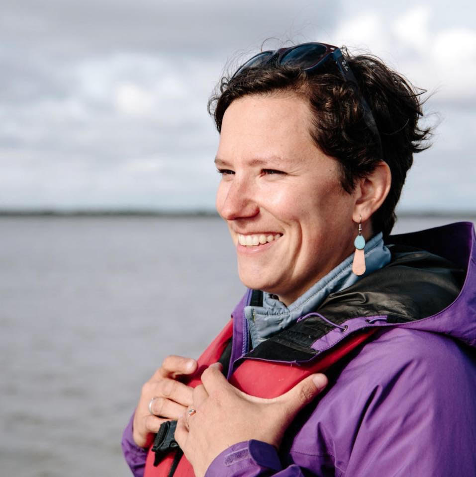 She is wearing a life jacket over a hooded raincoat. A large body of gray water extends behind her.