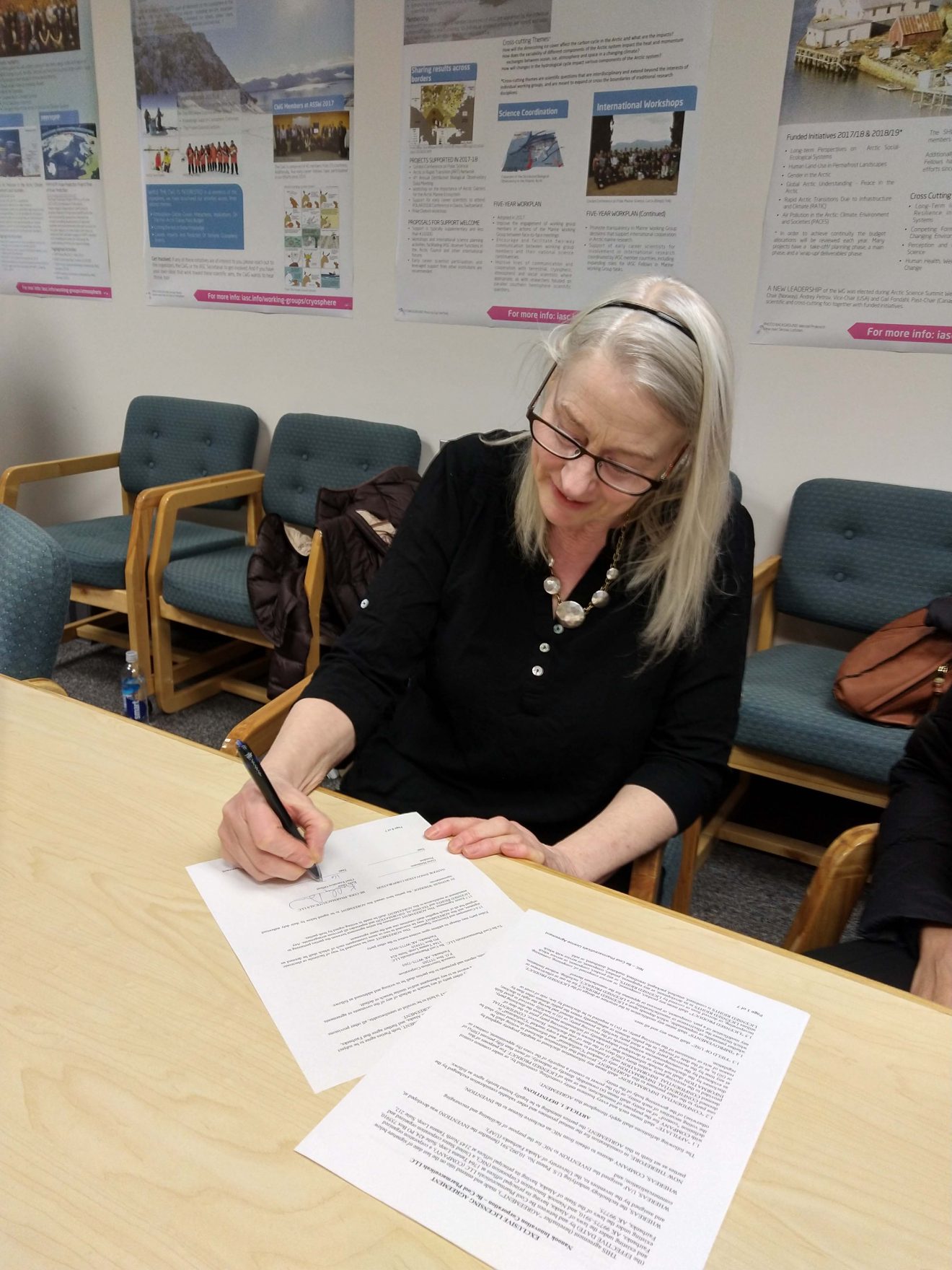 A woman signs a piece of paper in a conference room. Science posters hang on the wall behind her.