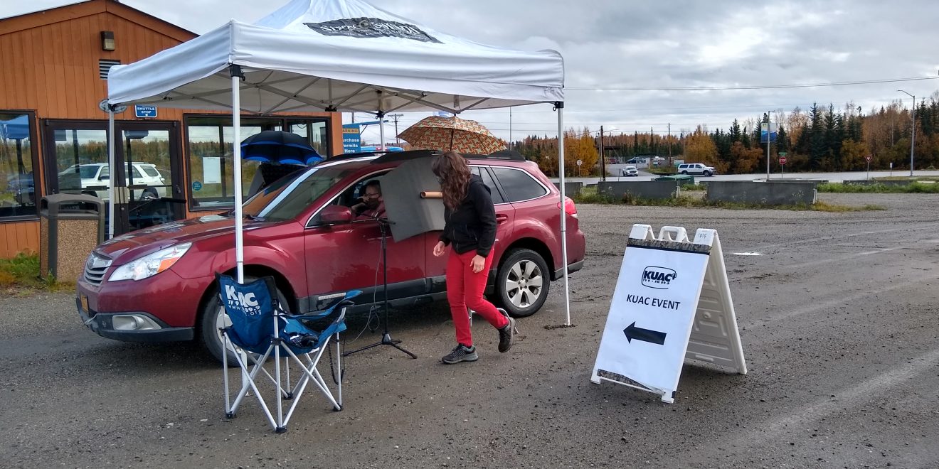 A car parks under a temporary awning. The driver is wearing a mask. Someone stands outside the car door, which is partially covered by a shield. A KUAC chair and sign are visible. They are in a parking lot.