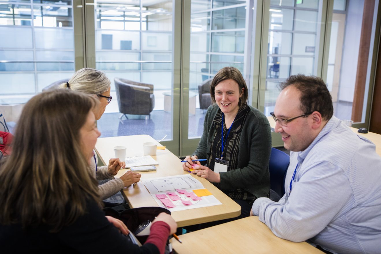 Four people sit around small desks. Papers and coffee cups are in front of them.