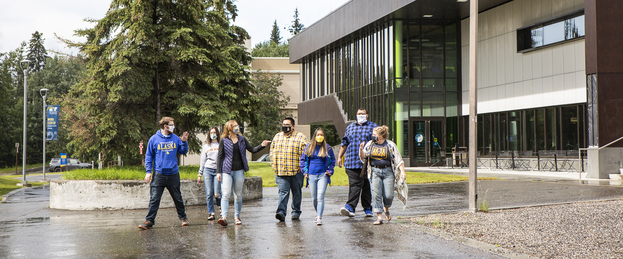 A group of seven students, all wearing masks, walk near Wood Center and the dining facility. It has been raining.