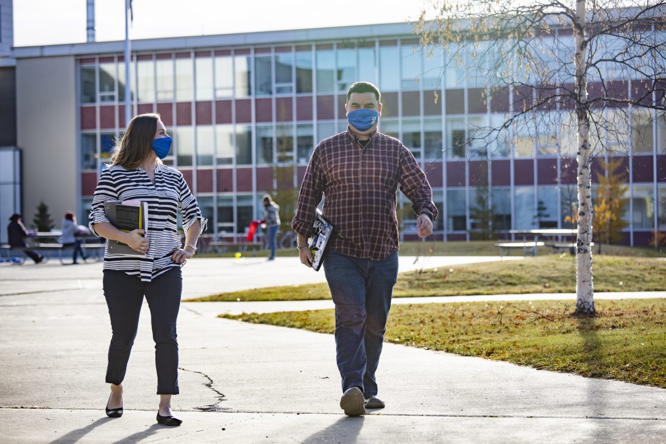 A woman and man, both wearing masks and carrying books, walk away from the Duckering Building. It is late fall. They are wearing civilian clothes.