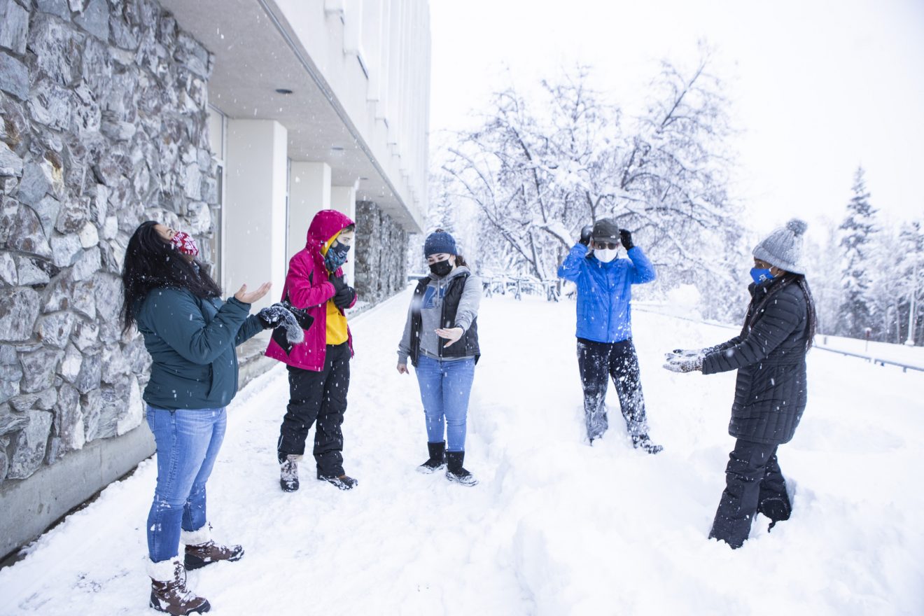 A group of students physically distanced and wearing masks. Snow is falling and they are standing in a lot of snow.