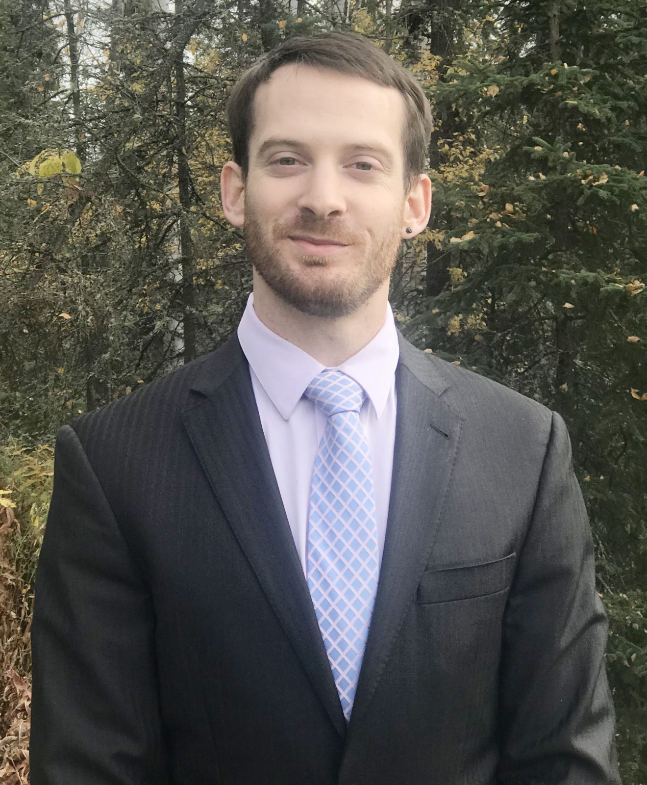 Head shot of a man in a suit standing outside. It is fall.