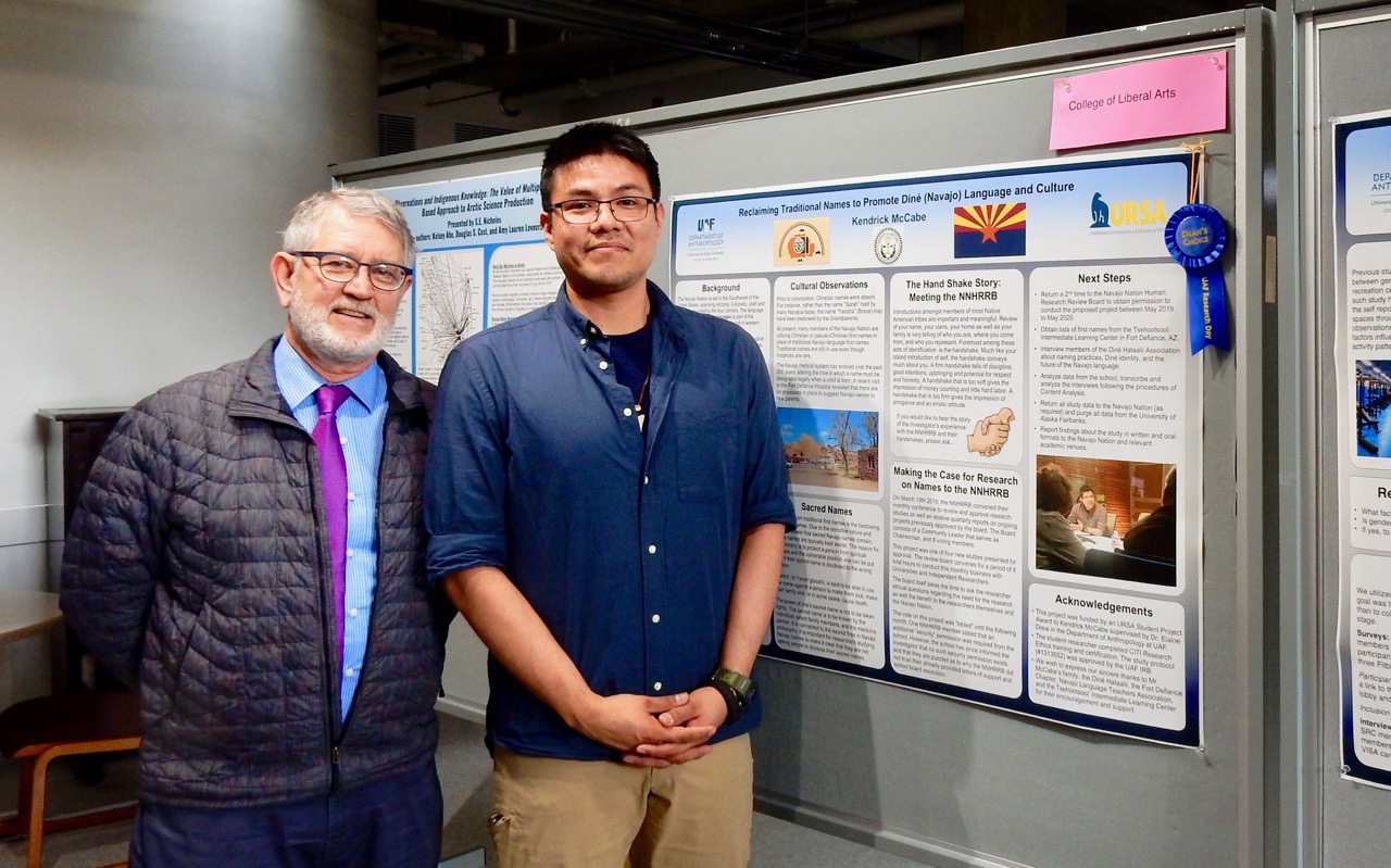 Two men stand beside a display board with posters on it. One of the posters has a blue ribbon pinned by it.