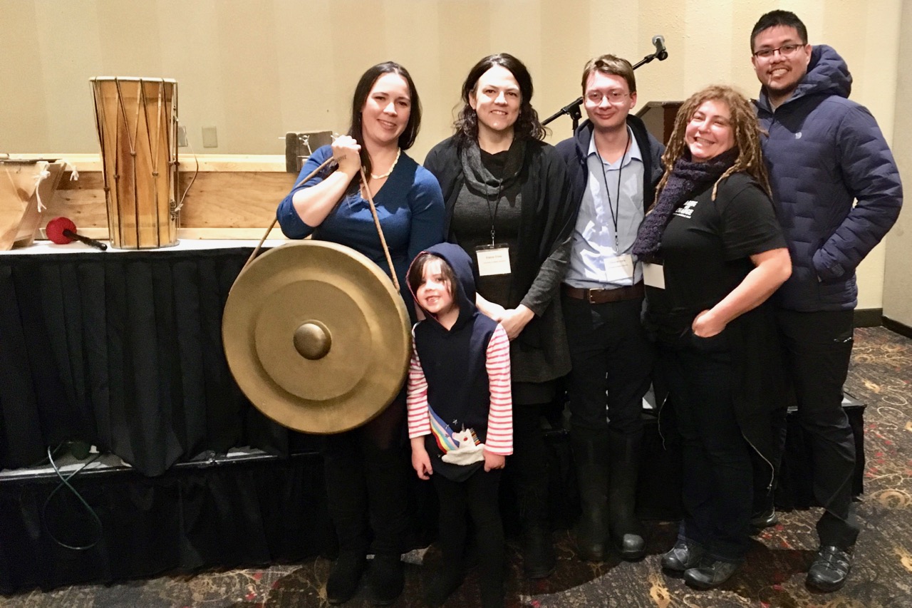 Five adults and a child pose for a photo in a large meeting room. One woman is holding what appears to be a shield.