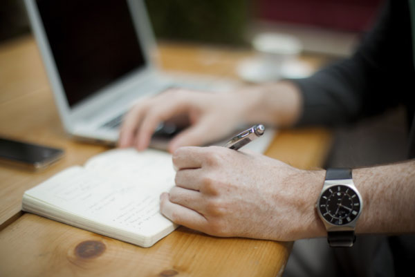 Left hand with watch on wrist writing notes with a laptop in the background.