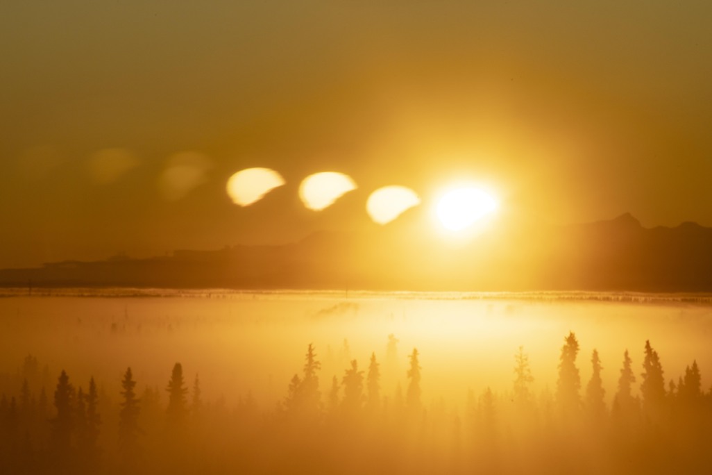 The sun peeks from behind the mountains. Multiple suns are reflected in the panes of glass. Ice fog blankets the valley, with trees in the foreground.
