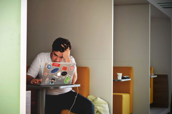 Young man looking at a laptop covered in stickers and running his fingers through his hair.